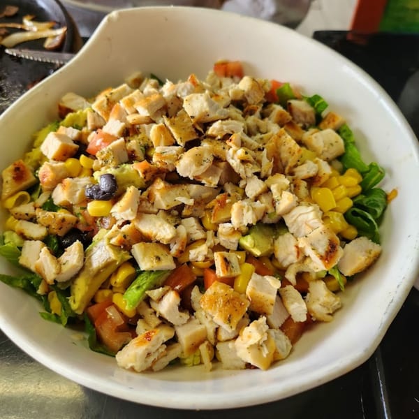 A white bowl filled with a vibrant salad of grilled chicken, corn, avocado, tomatoes, and lettuce.