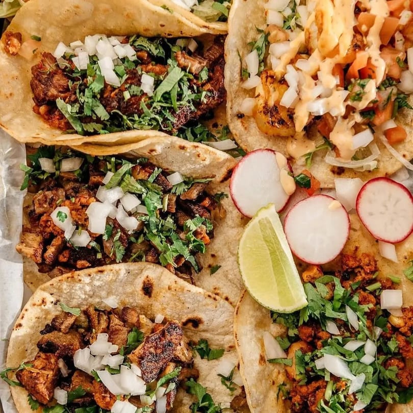 Close-up of several Guac Tacos, garnished with cilantro, diced onions, radishes, and lime wedges.