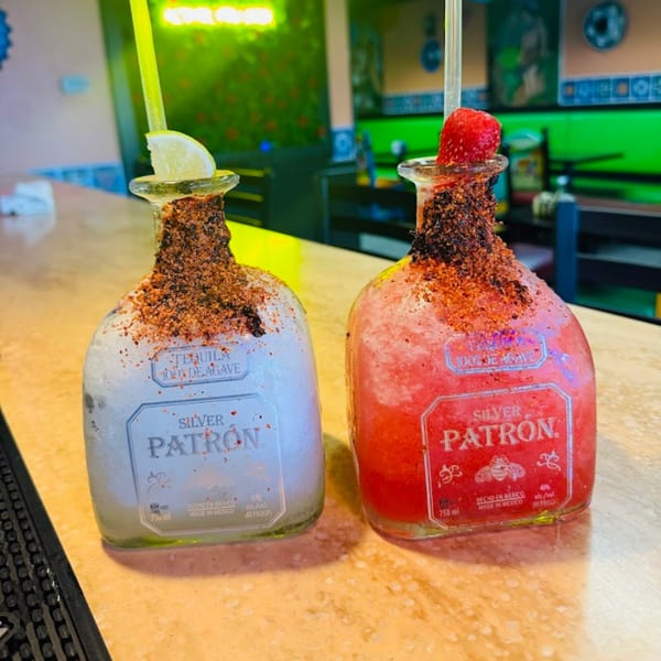 Two Patron tequila bottles, one white and one red, filled with frozen margaritas on a bar.