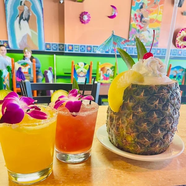 Three vibrant Guac Taco & Tequila Bar drinks, including a pineapple cocktail, on a table with colorful decor.