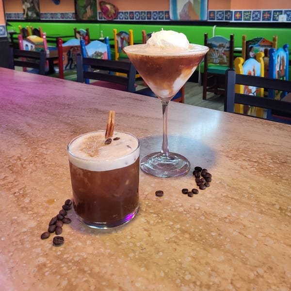 Two coffee cocktails on a bar, one with whipped cream, against a colorful restaurant background.
