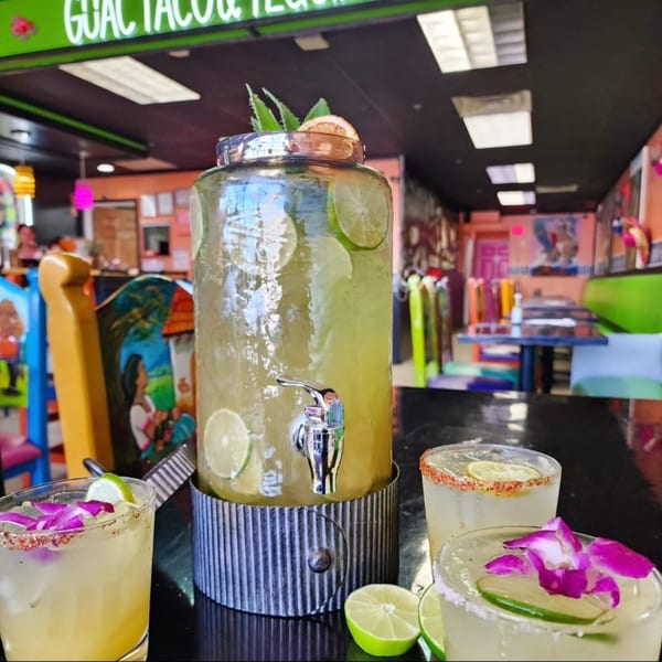 A large drink dispenser and three margaritas with lime and flower garnishes at Guac Taco & Tequila Bar.