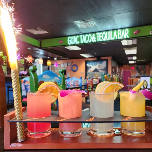 A flight of colorful margaritas with fruit garnishes and a sparkler, inside Guac Taco & Tequila Bar.